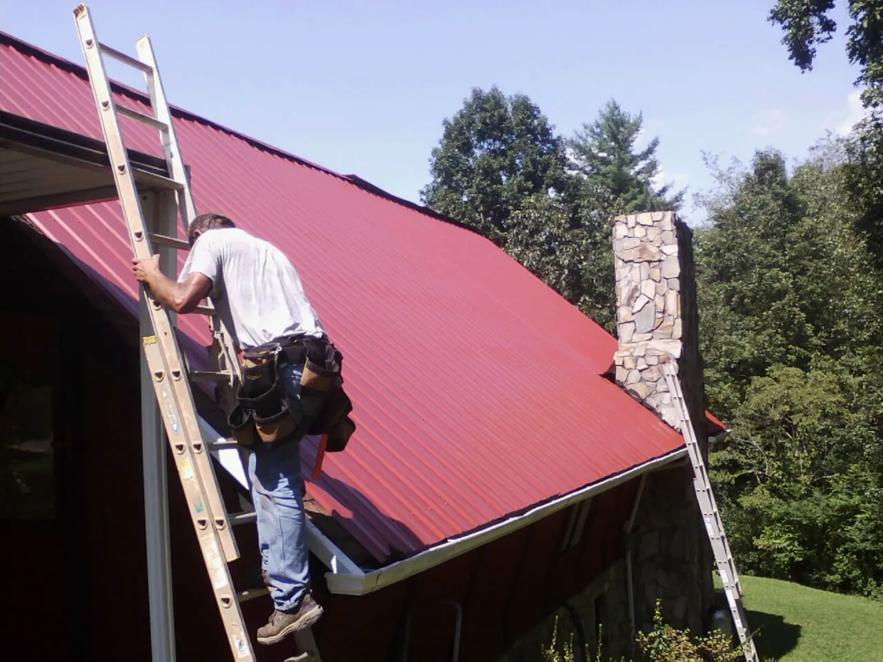 Roofer on a ladder beside a red metal roof and stone chimney — Soffit & Fascia Repair by Roofing Brothers in Waxahachie